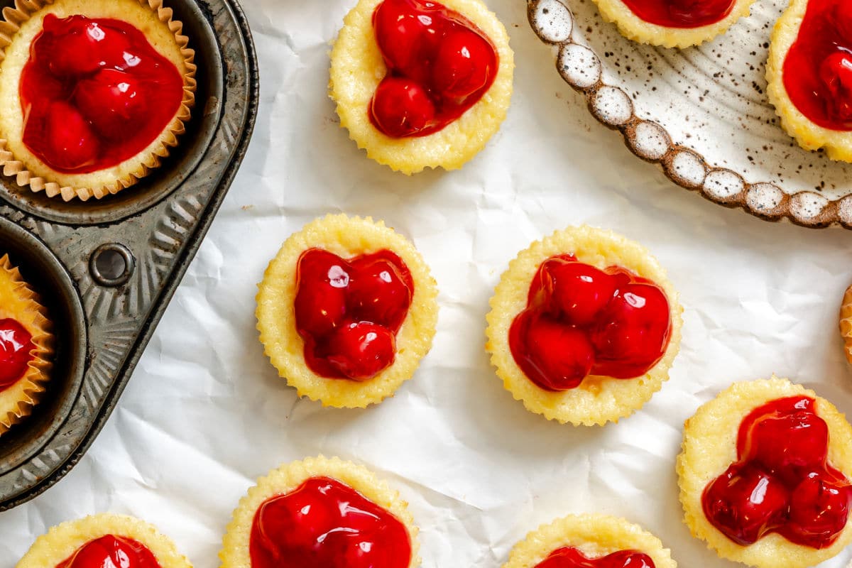 Mini cheesecakes topped with bright red cherry filling on parchment paper, with a muffin tin on the side.