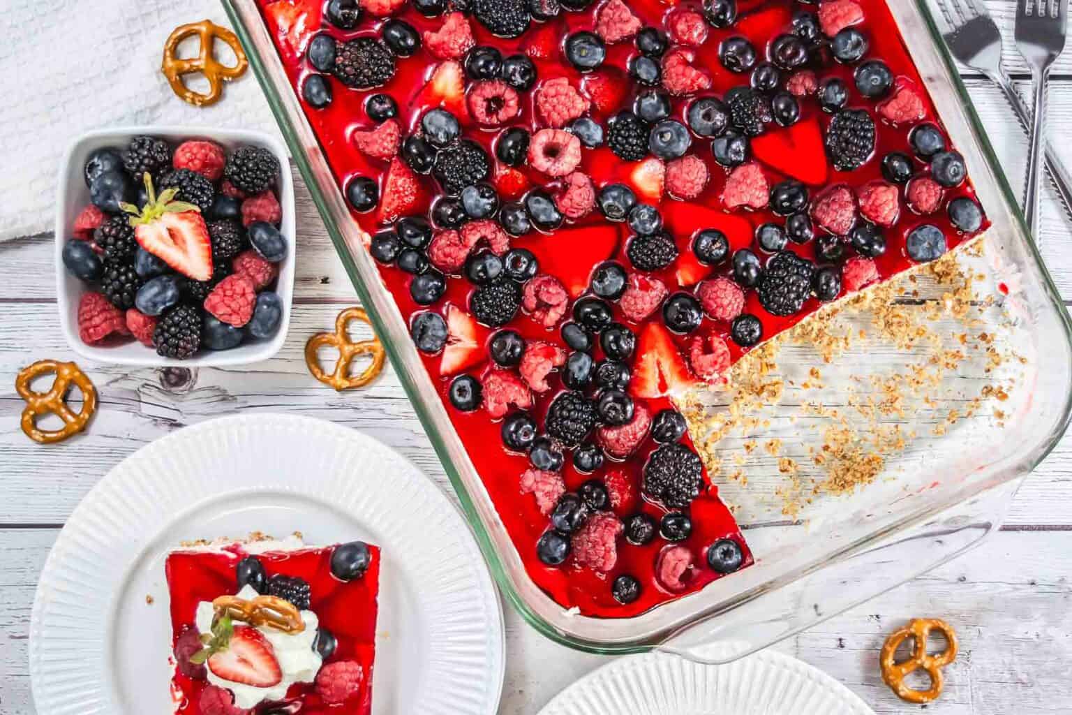 A berry-topped gelatin dessert in a glass dish, served with whipped cream and pretzels on white plates.
