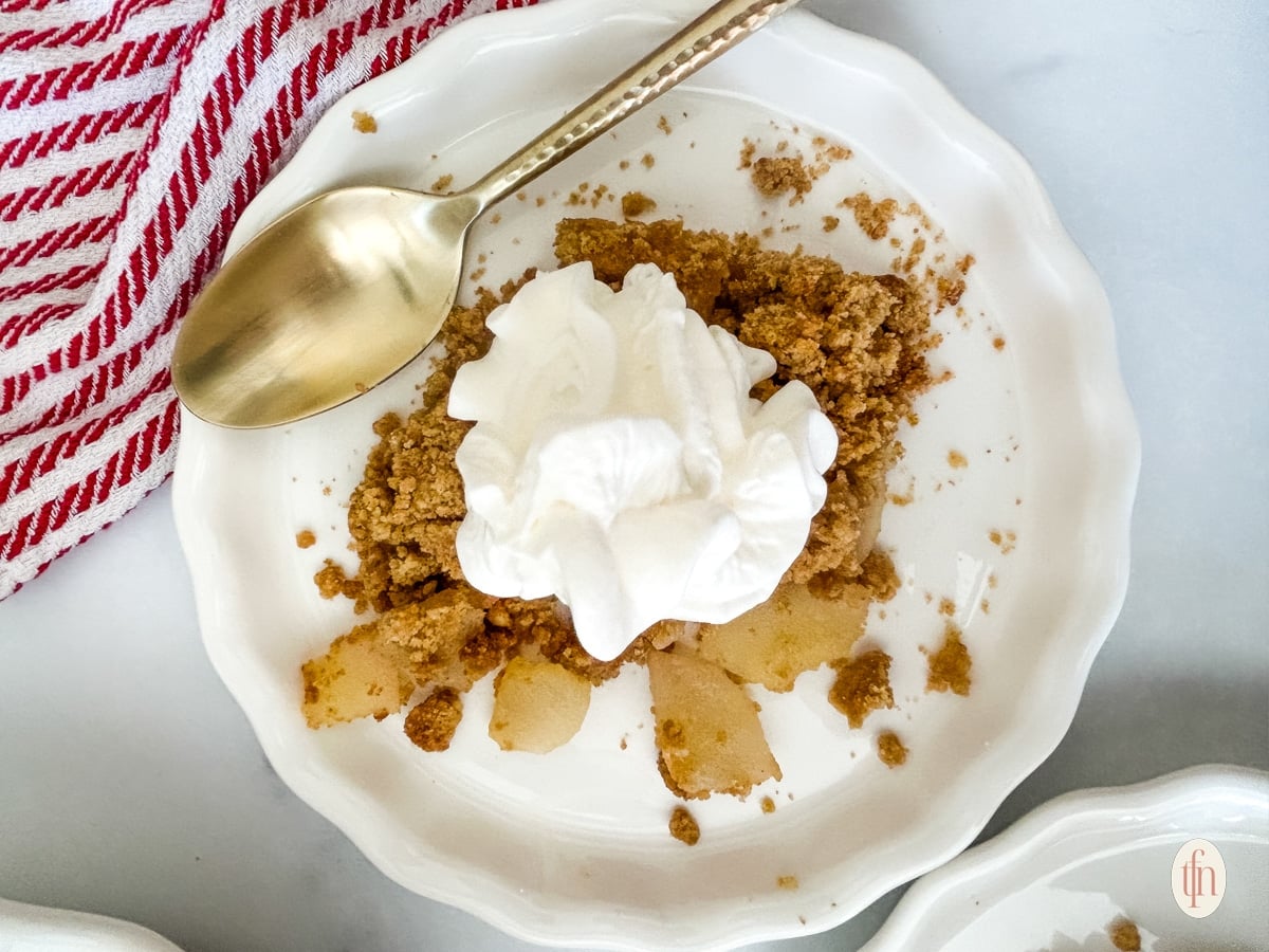 A plate of apple crisp topped with whipped cream and a gold spoon on the side.