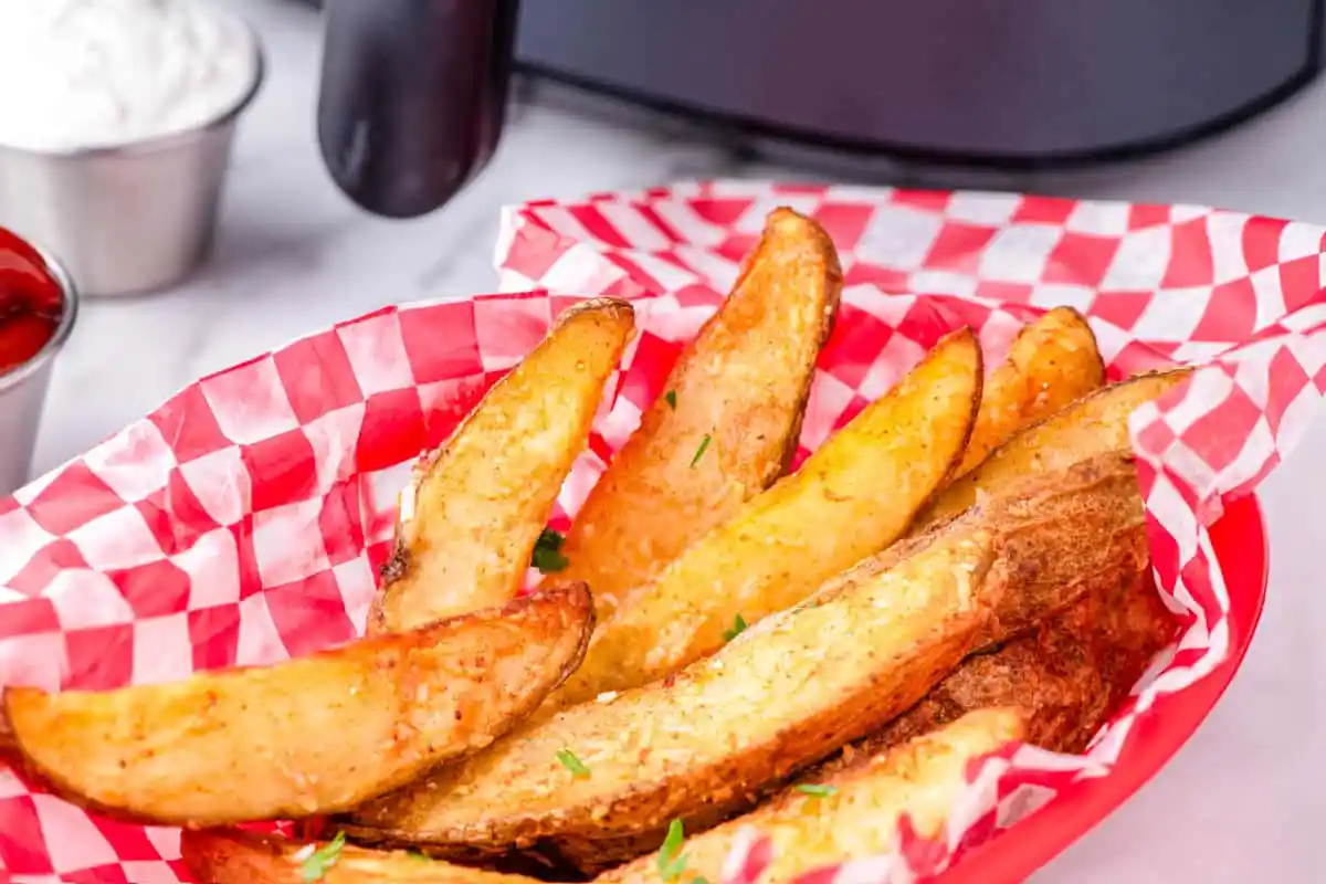 Golden potato wedges in a red basket with red checkered paper, served with dips in the background.