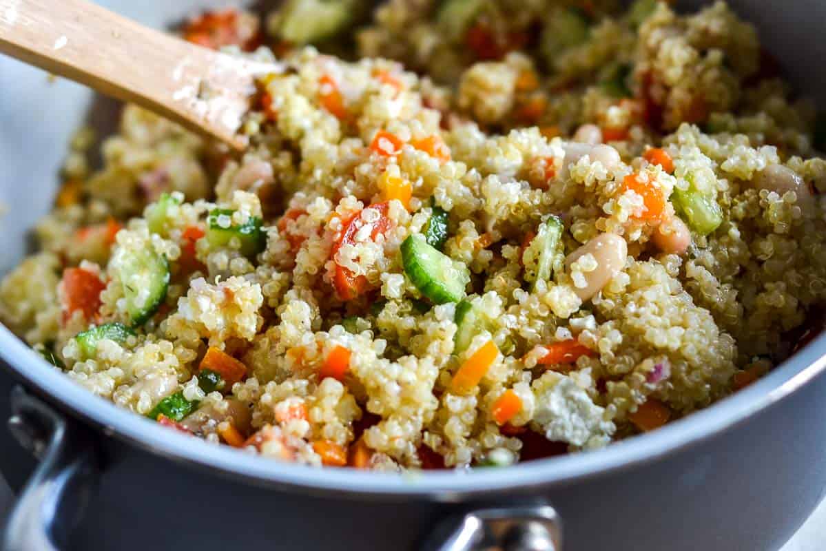 A close-up of quinoa salad with chopped vegetables being stirred with a wooden spoon in a pot.