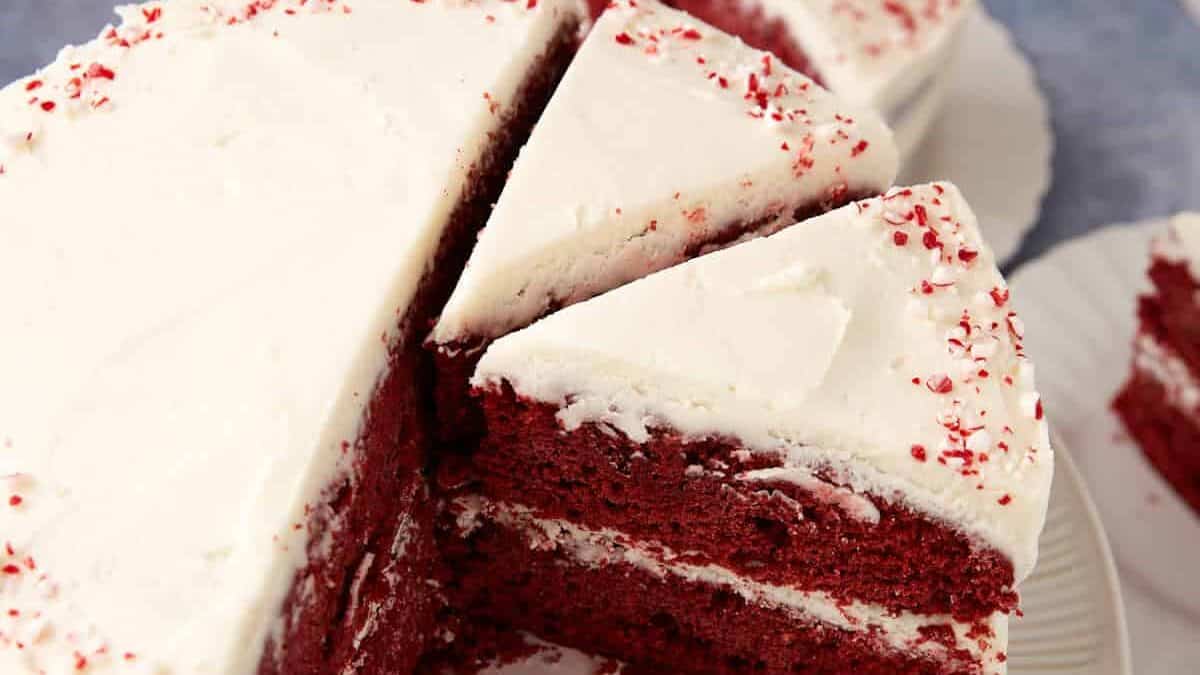 A sliced red velvet cake with white frosting and red sprinkles on top, shown on a plate.