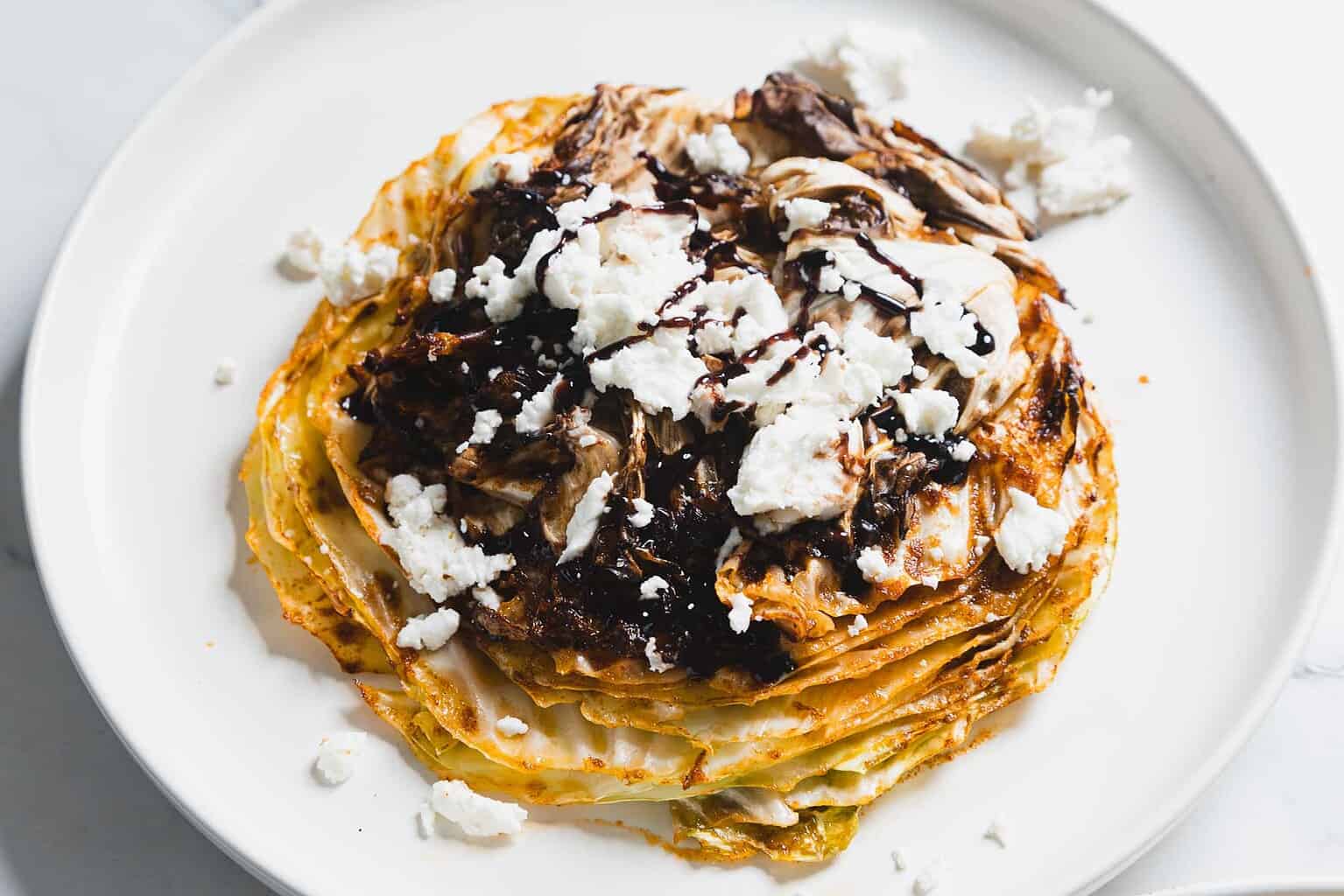Grilled cabbage steak topped with crumbled cheese and a drizzle of balsamic glaze on a white plate.