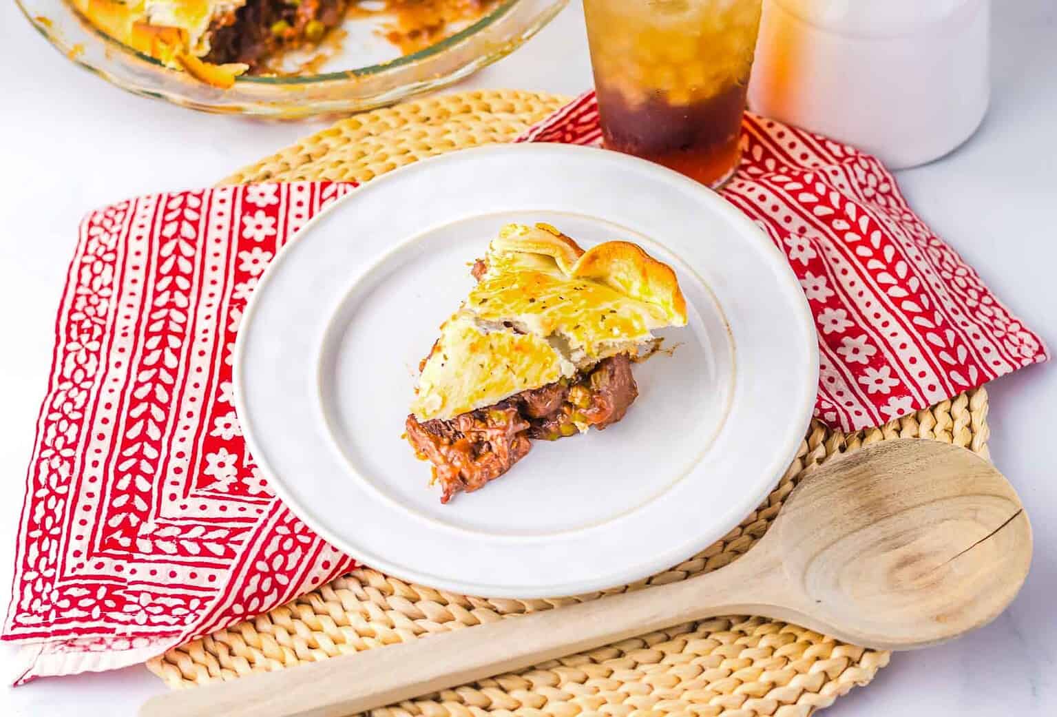 A slice of meat pie on a white plate with a red patterned napkin, wooden spoon, and iced drink nearby.