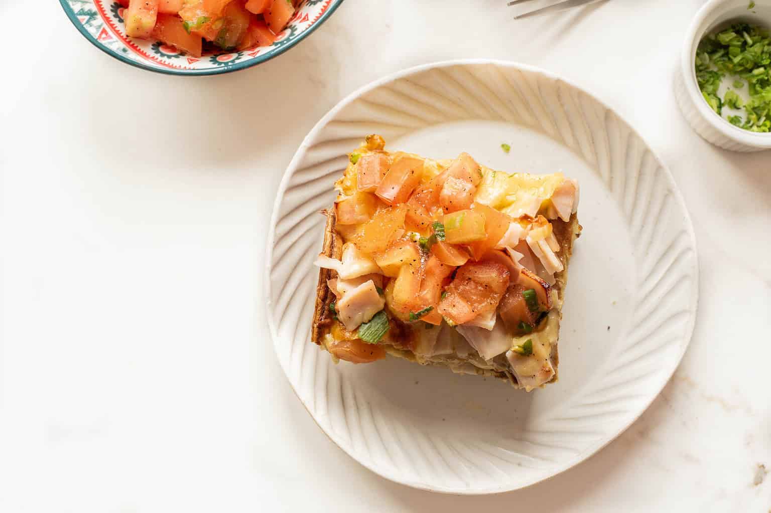 A slice of casserole topped with chopped tomatoes and herbs on a white plate.