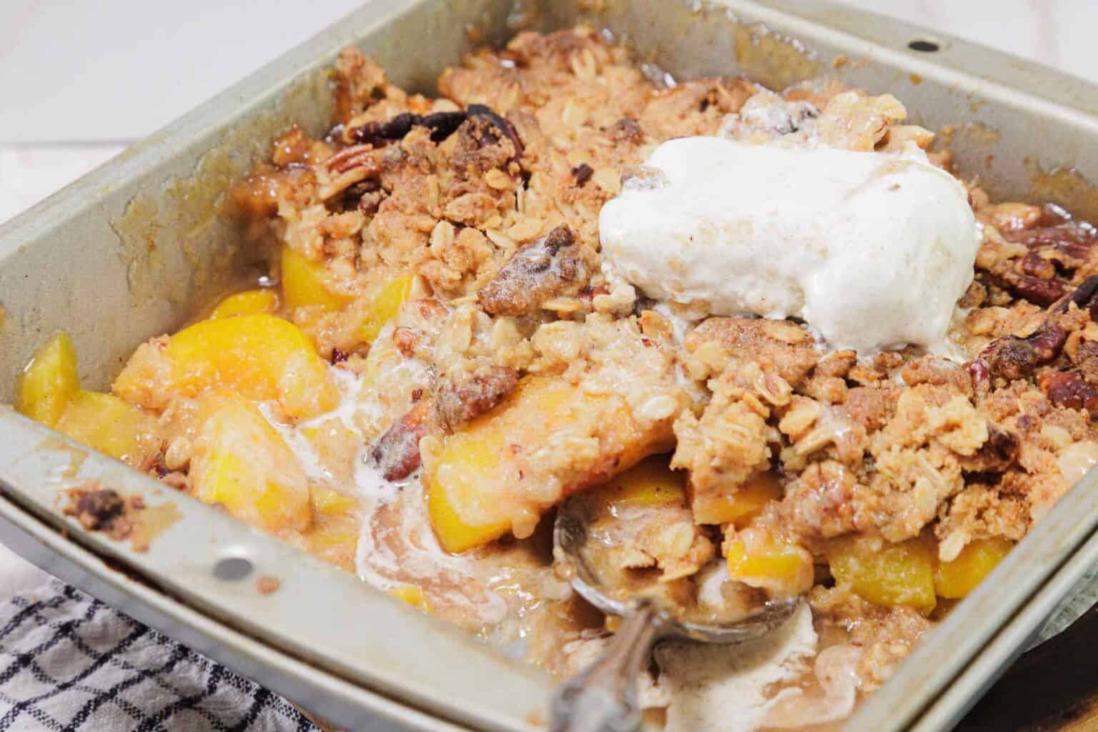 Peach crisp with oat topping and a dollop of whipped cream in a baking pan with a serving spoon.