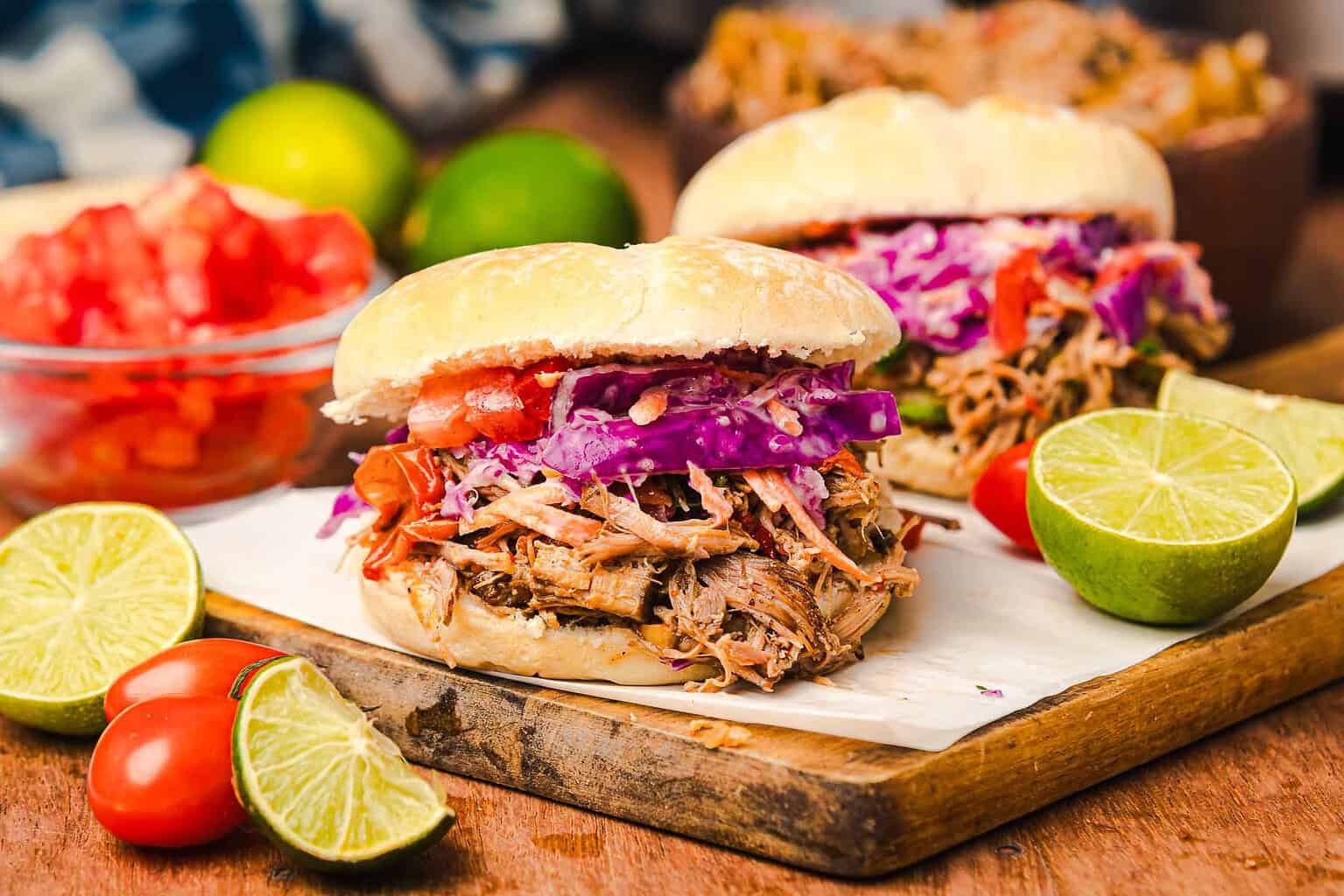 Pulled pork sandwiches topped with colorful slaw, surrounded by limes, tomatoes, and a bowl of chopped tomatoes.