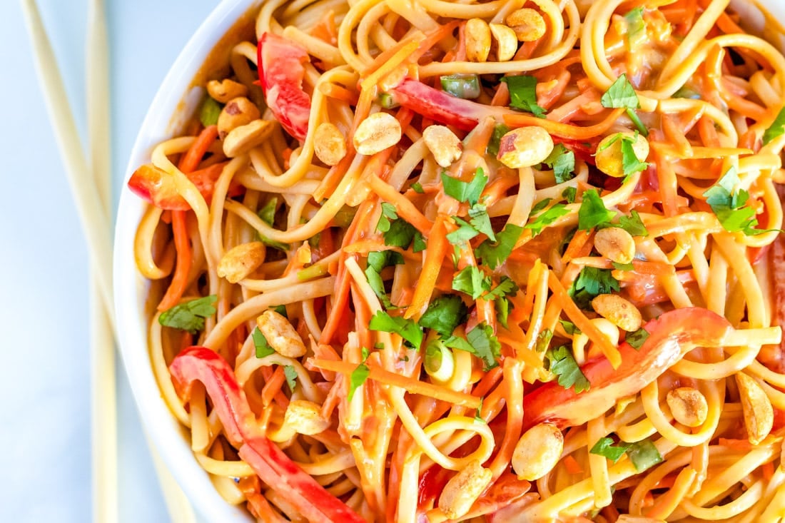 Bowl of noodle salad with red bell peppers, carrots, peanuts, and cilantro, with chopsticks beside it.