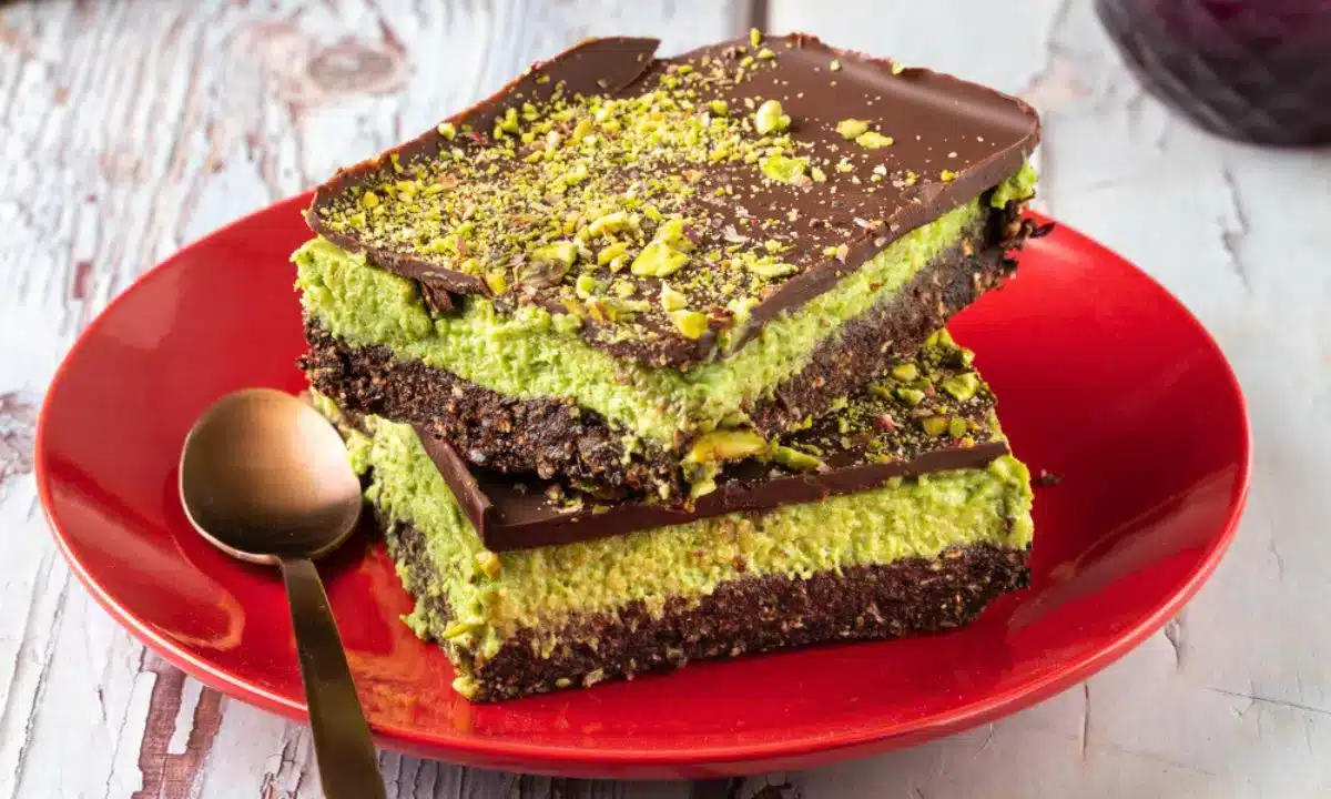 Two chocolate and pistachio dessert bars stacked on a red plate with a spoon beside them.