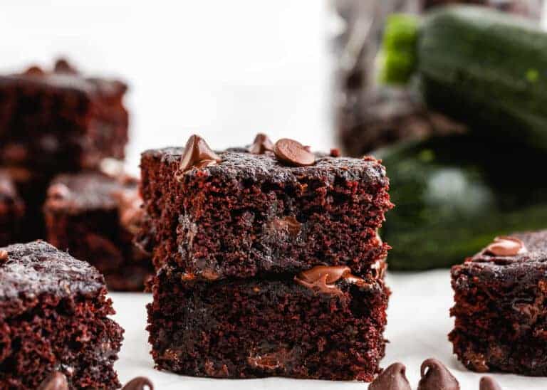 Two chocolate brownies stacked, topped with chocolate chips, with more brownies and zucchini in the background.