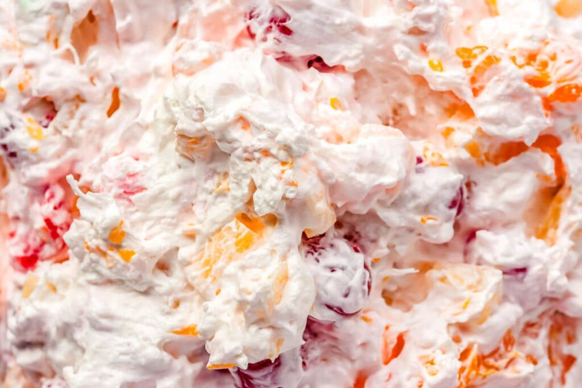 Close-up of a creamy fruit salad with whipped topping, featuring pieces of orange, red, and purple fruit.