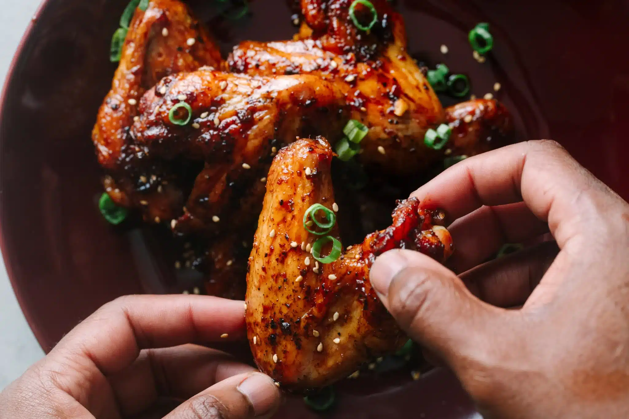 Hands holding a glazed chicken wing garnished with green onions and sesame seeds on a dark plate.