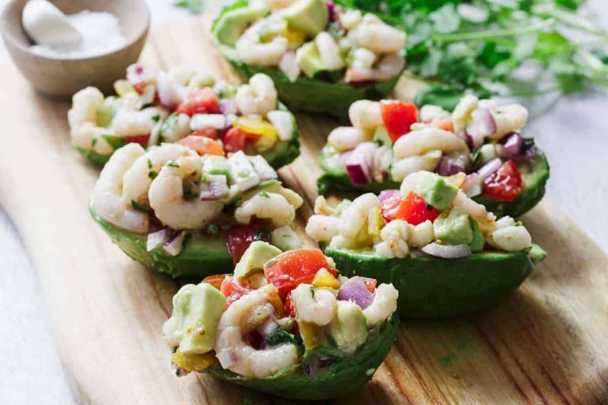 Avocado halves filled with shrimp ceviche, diced tomatoes, red onion, and cilantro on a wooden board.