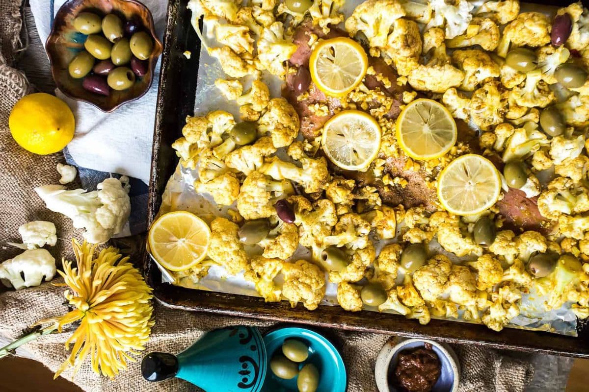Roasted cauliflower with olives and lemon slices on a baking sheet, surrounded by olives, flowers, and condiments.