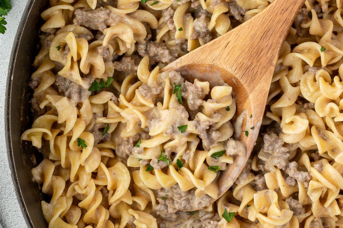 A wooden spoon scoops creamy beef and noodles garnished with parsley from a skillet.