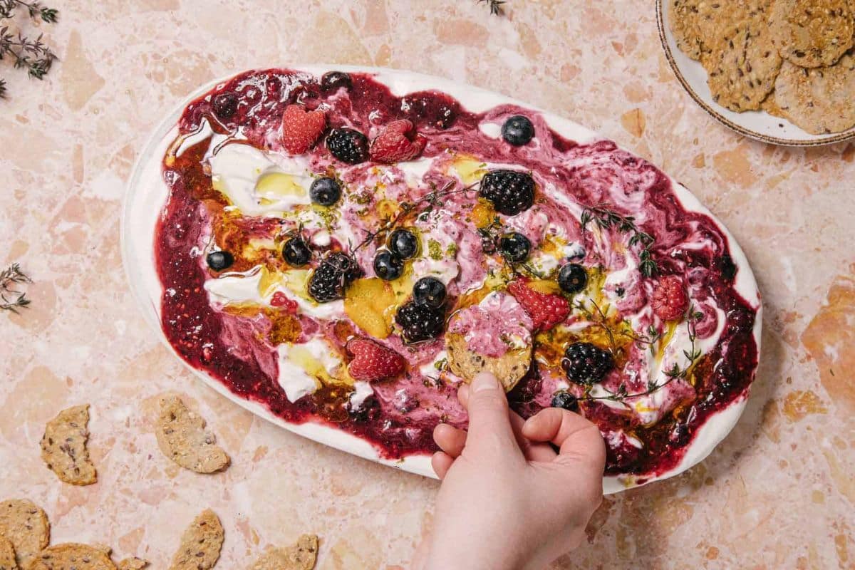 A hand dips a cracker into a colorful yogurt and berry swirl dip on an oval platter.