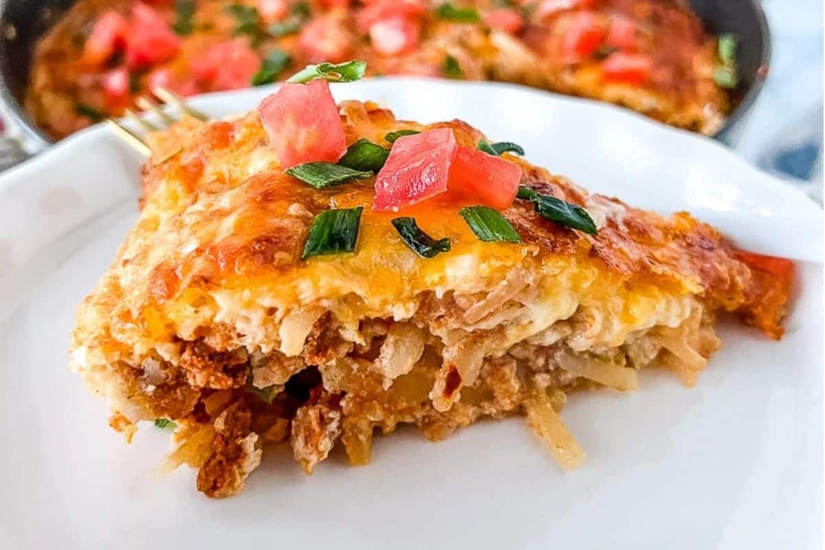 A slice of cheesy casserole topped with chopped tomatoes and green onions on a white plate.
