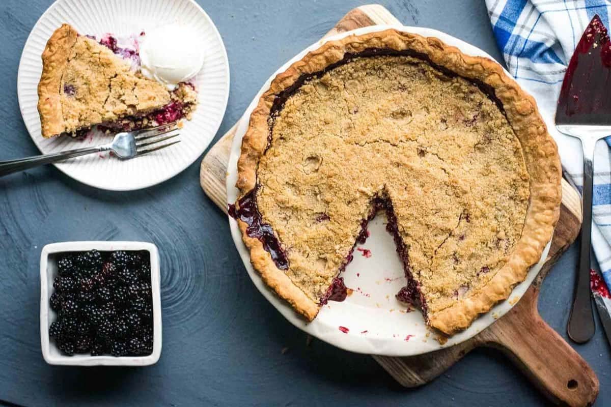 Whole blackberry pie with a slice missing, served with ice cream. Fresh blackberries in a bowl nearby.