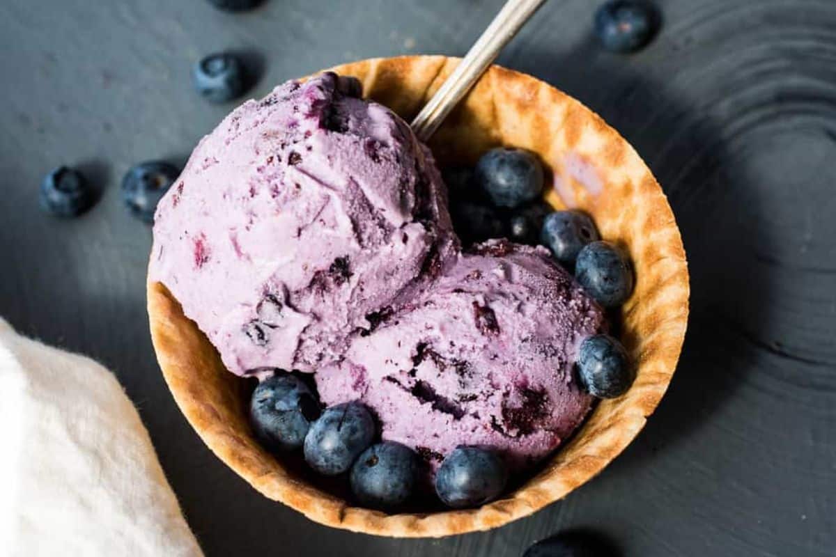 Two scoops of blueberry ice cream in a waffle bowl, garnished with fresh blueberries and a spoon.
