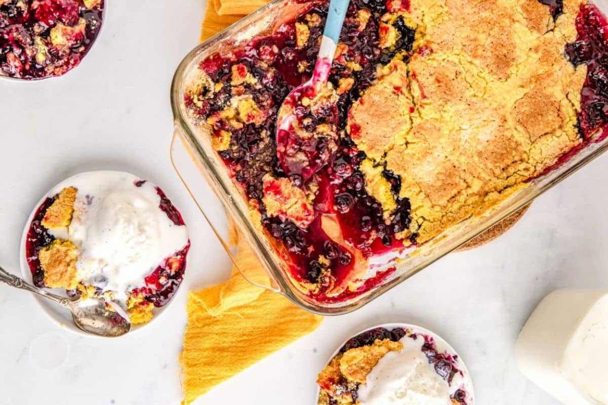 A baking dish of berry cobbler with plates of cobbler and vanilla ice cream on a white table.