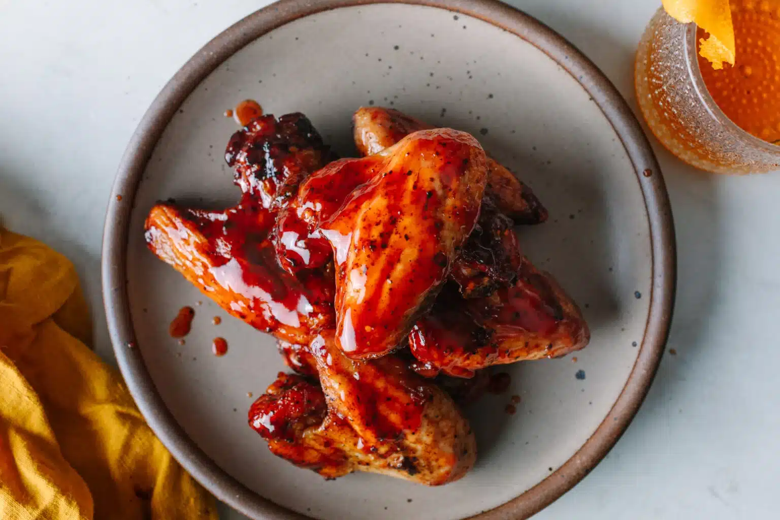 A plate of glazed chicken wings with a shiny, reddish sauce, next to a drink and a yellow napkin.