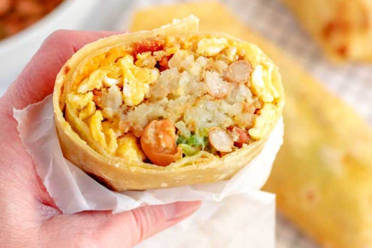 Hand holding a breakfast burrito cut in half, showing eggs, beans, potatoes, and cheese inside.