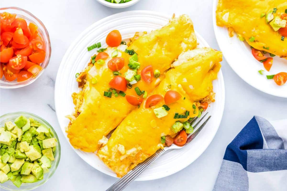 Two cheesy enchiladas topped with chopped tomatoes, avocado, and green onions on a white plate with a fork.