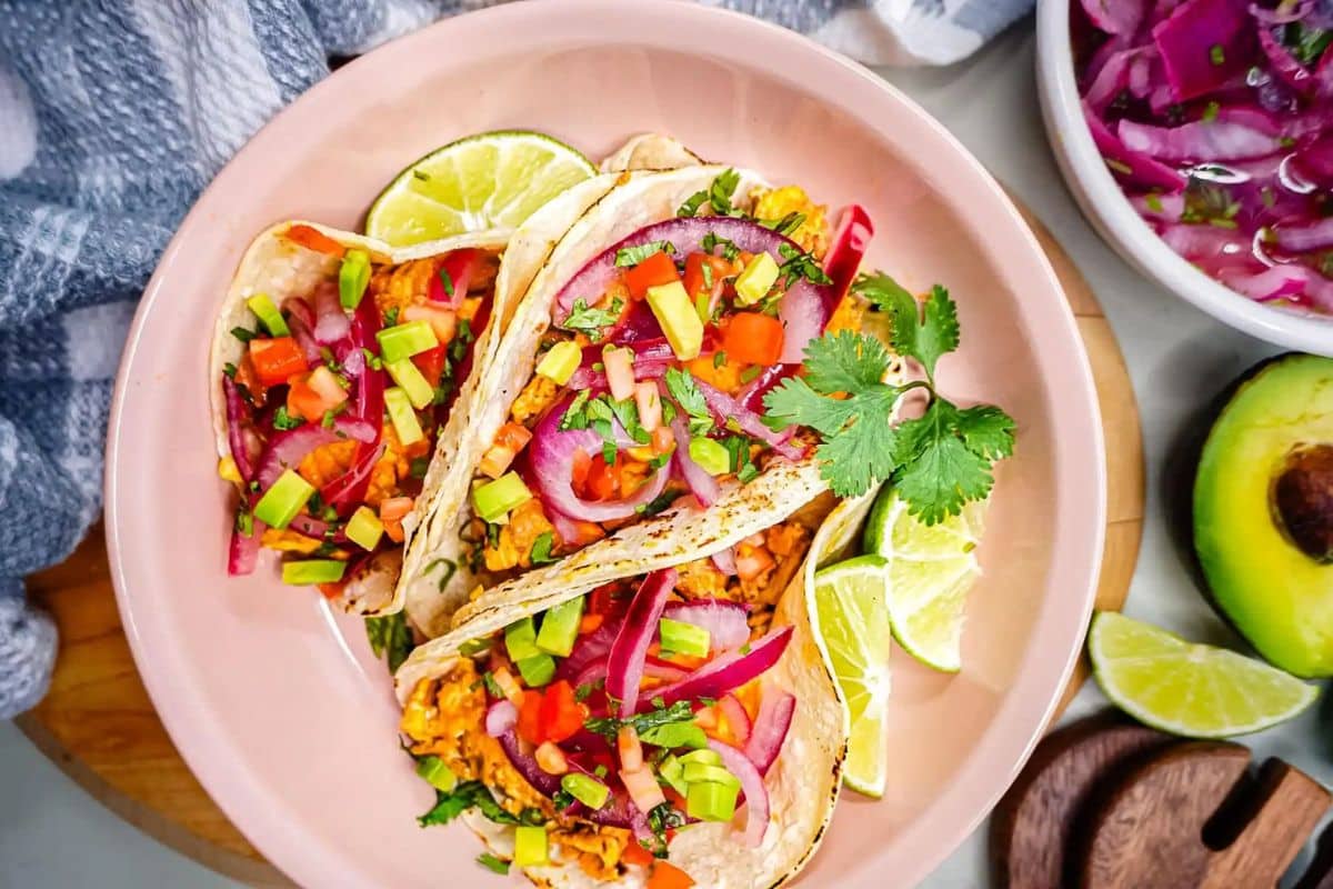 Three colorful tacos with veggies and pickled onions on a plate, garnished with lime wedges and cilantro.