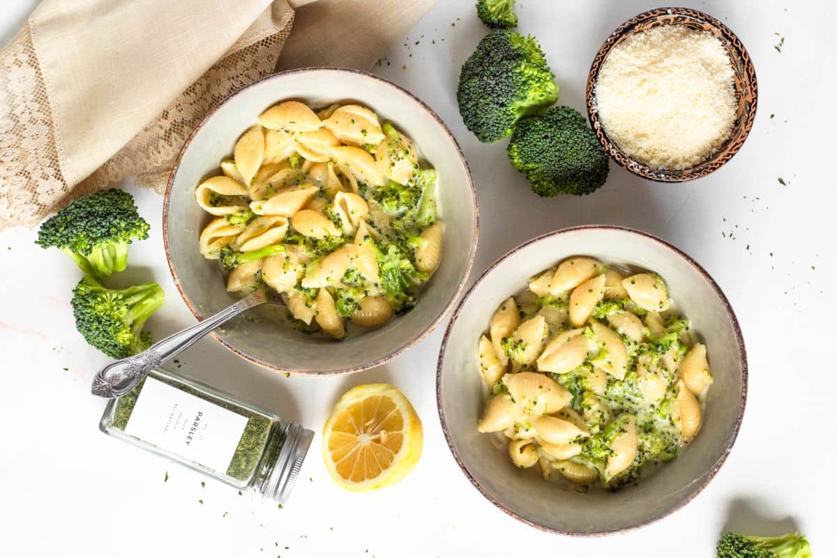 Two bowls of pasta with broccoli and cheese, surrounded by broccoli, lemon, grated cheese, and spices on a table.