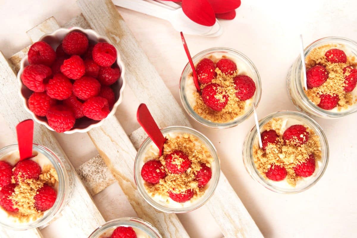 Six glass cups with layered dessert topped with raspberries and crumbs, next to a bowl of fresh raspberries.