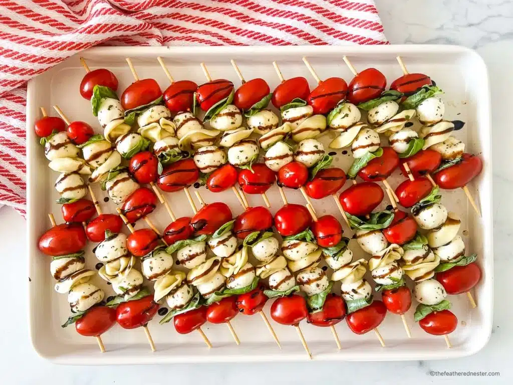 Skewers with tomatoes, mozzarella, basil, and pasta drizzled with balsamic glaze on a white tray.