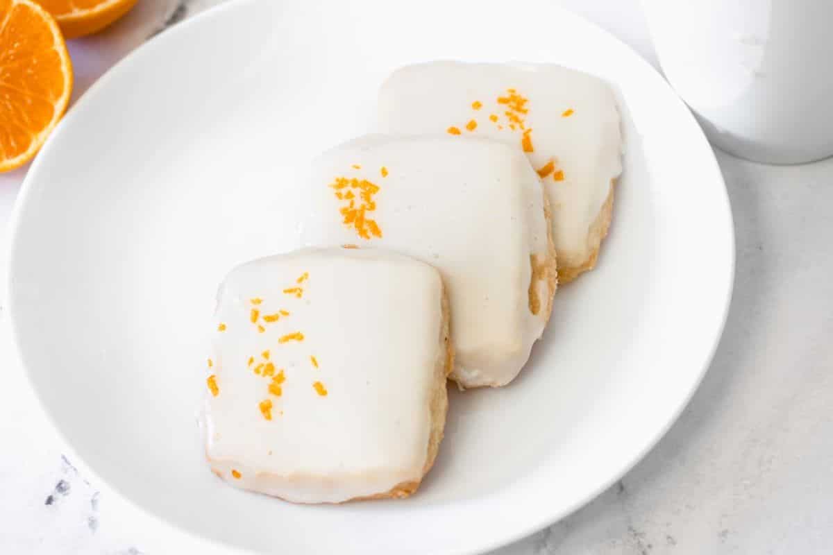 Three square cookies with white icing and orange zest on top, arranged on a white plate.