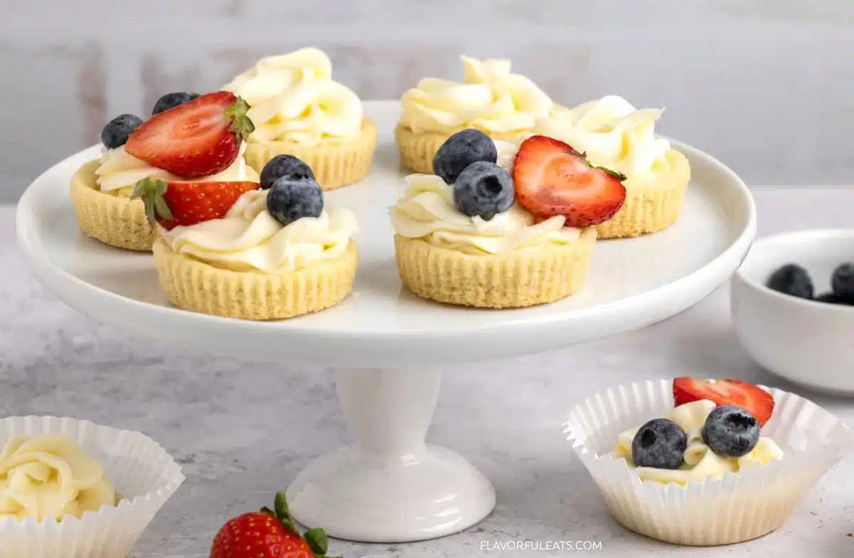 Mini cheesecakes with whipped cream, strawberries, and blueberries on a white cake stand, plus extra berries nearby.