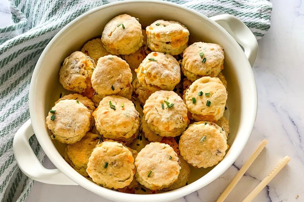 A white dish filled with fresh, round biscuits topped with chopped chives on a marble surface.