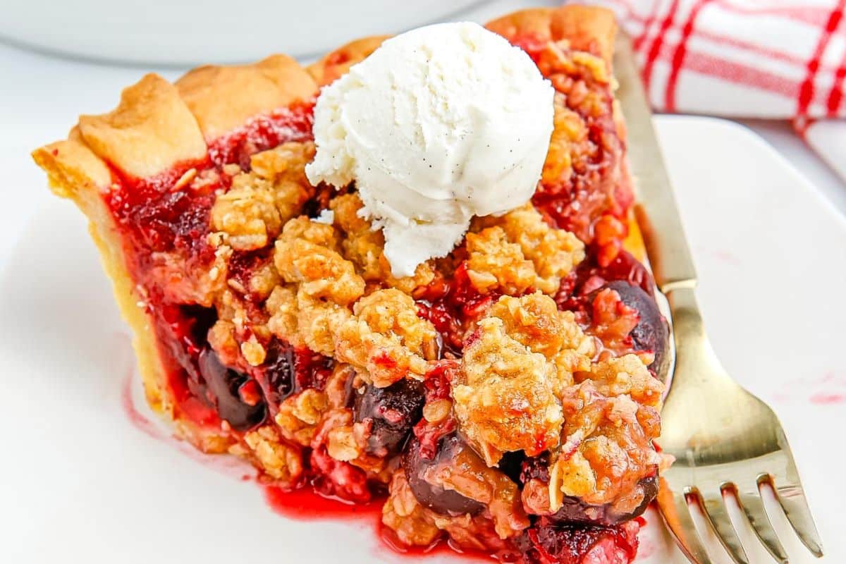 A slice of cherry crumble pie topped with a scoop of vanilla ice cream on a white plate with a gold fork.