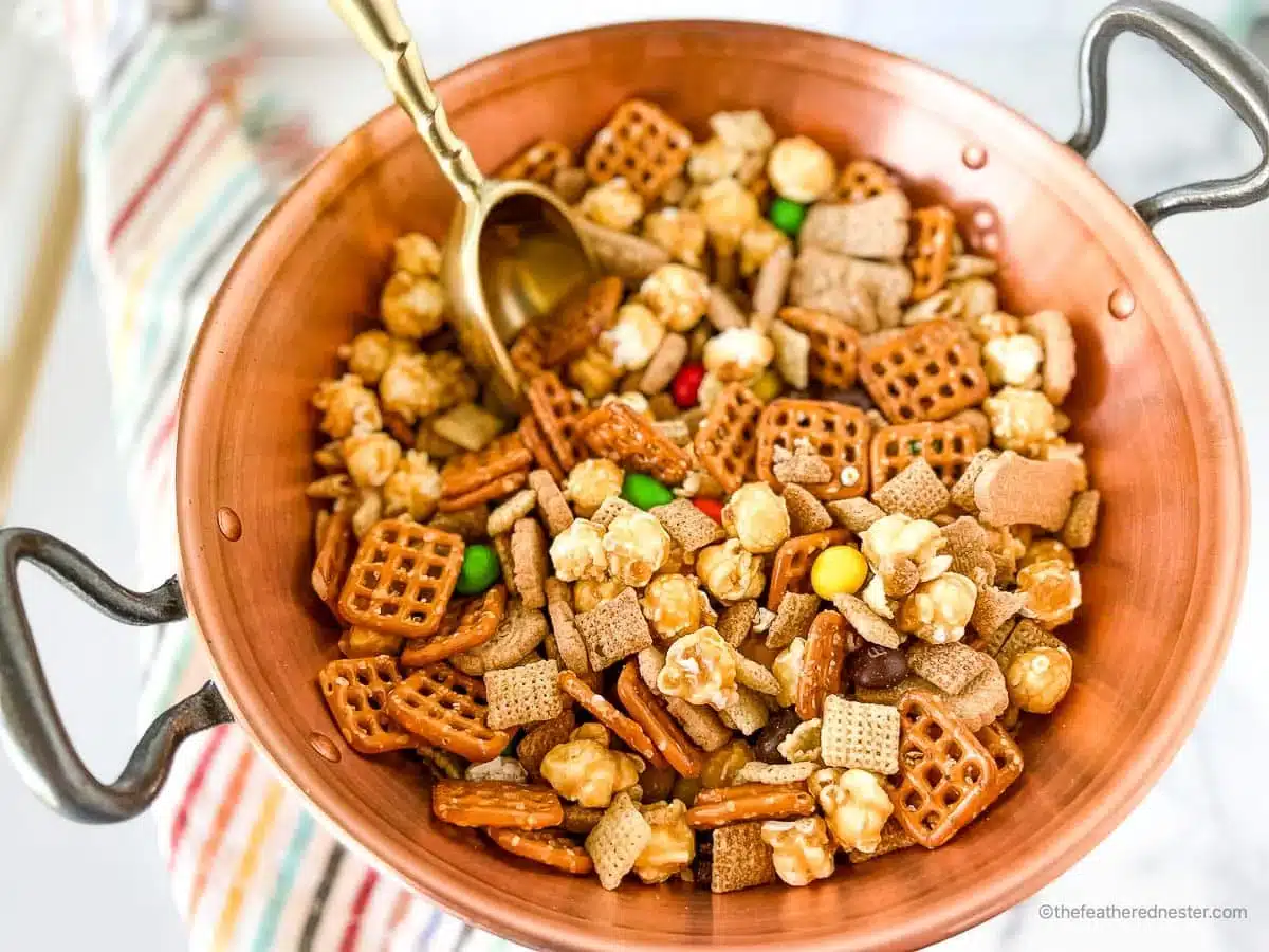 A copper bowl filled with snack mix including pretzels, popcorn, cereal, and colorful candy pieces.