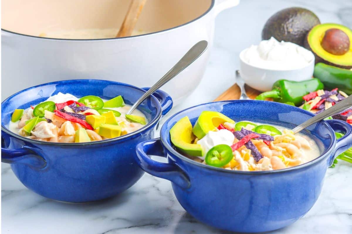 Two blue bowls of creamy soup topped with avocado, jalapeño, sour cream, and tortilla strips on a marble surface.