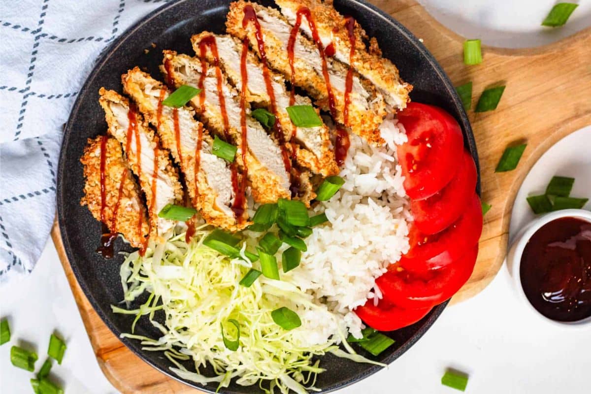 A black plate with sliced breaded chicken, white rice, shredded cabbage, tomato slices, and green onions.