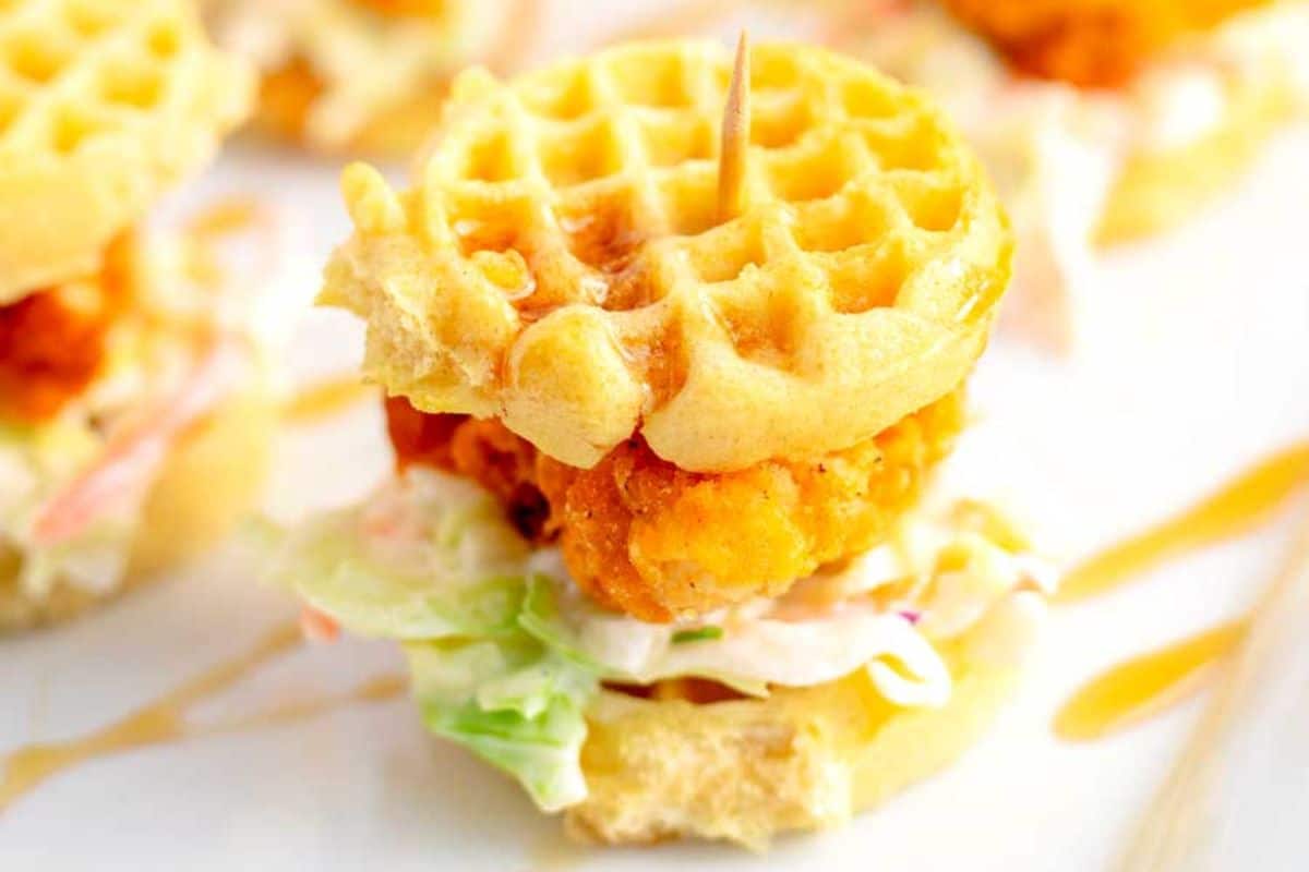 Mini waffle sandwich with fried chicken and coleslaw, secured with a toothpick, drizzled with syrup.