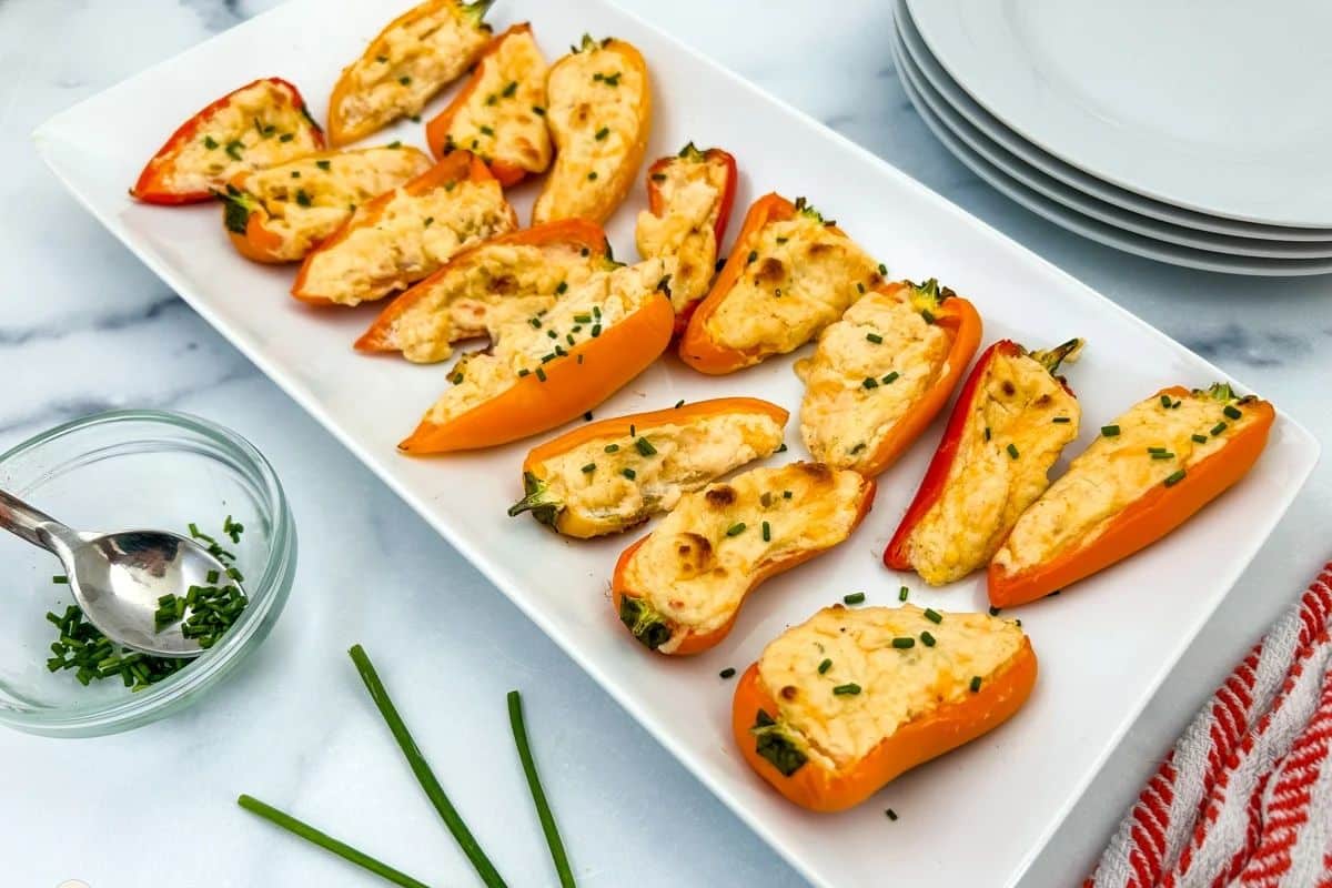 Mini stuffed peppers filled with melted cheese and herbs, arranged on a white platter with chopped chives.