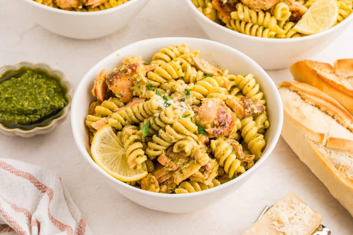 A bowl of pesto pasta with salmon, garnished with a lemon slice, next to bread and a small bowl of pesto.