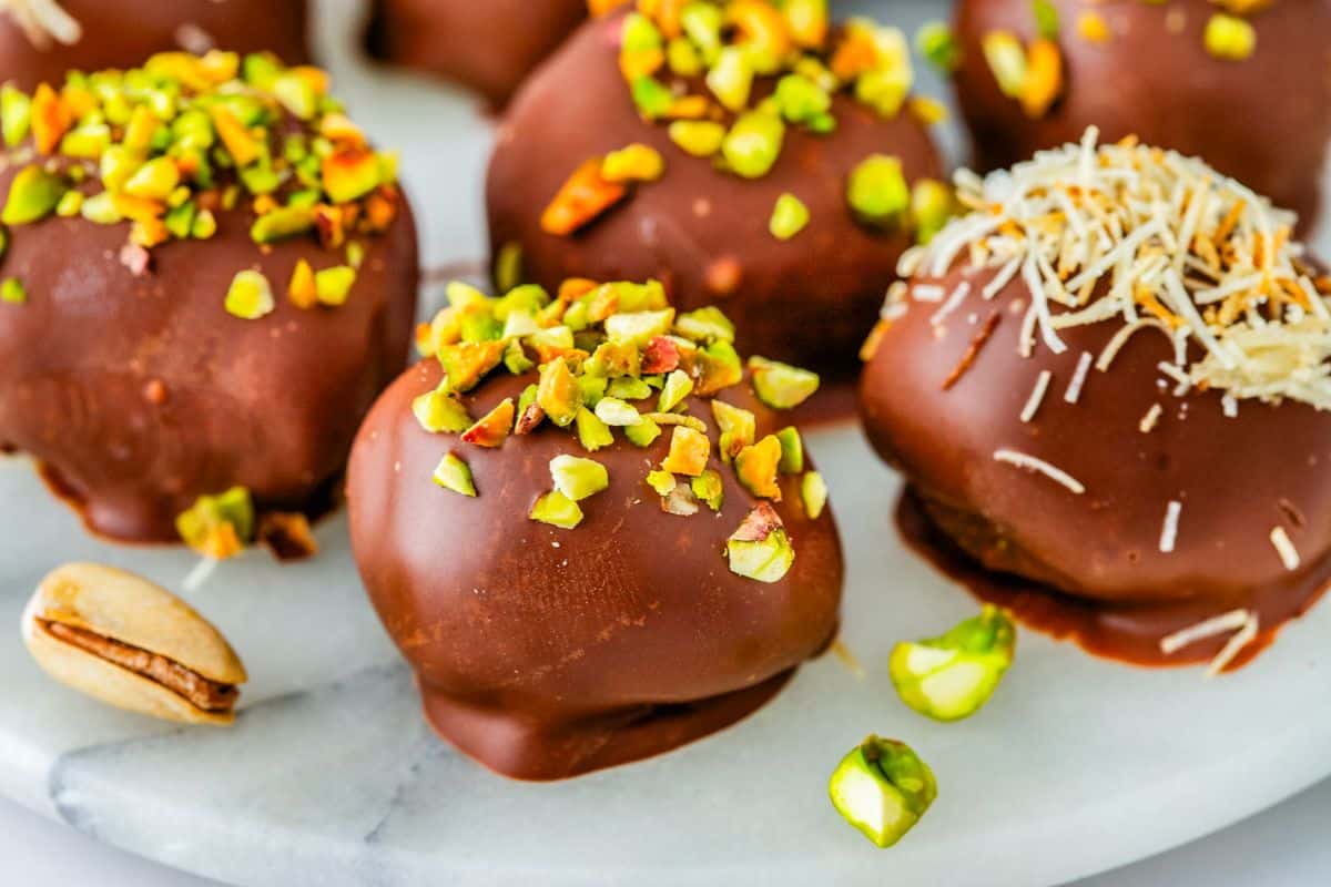 Chocolate-covered truffles topped with chopped pistachios and shredded coconut on a white surface.