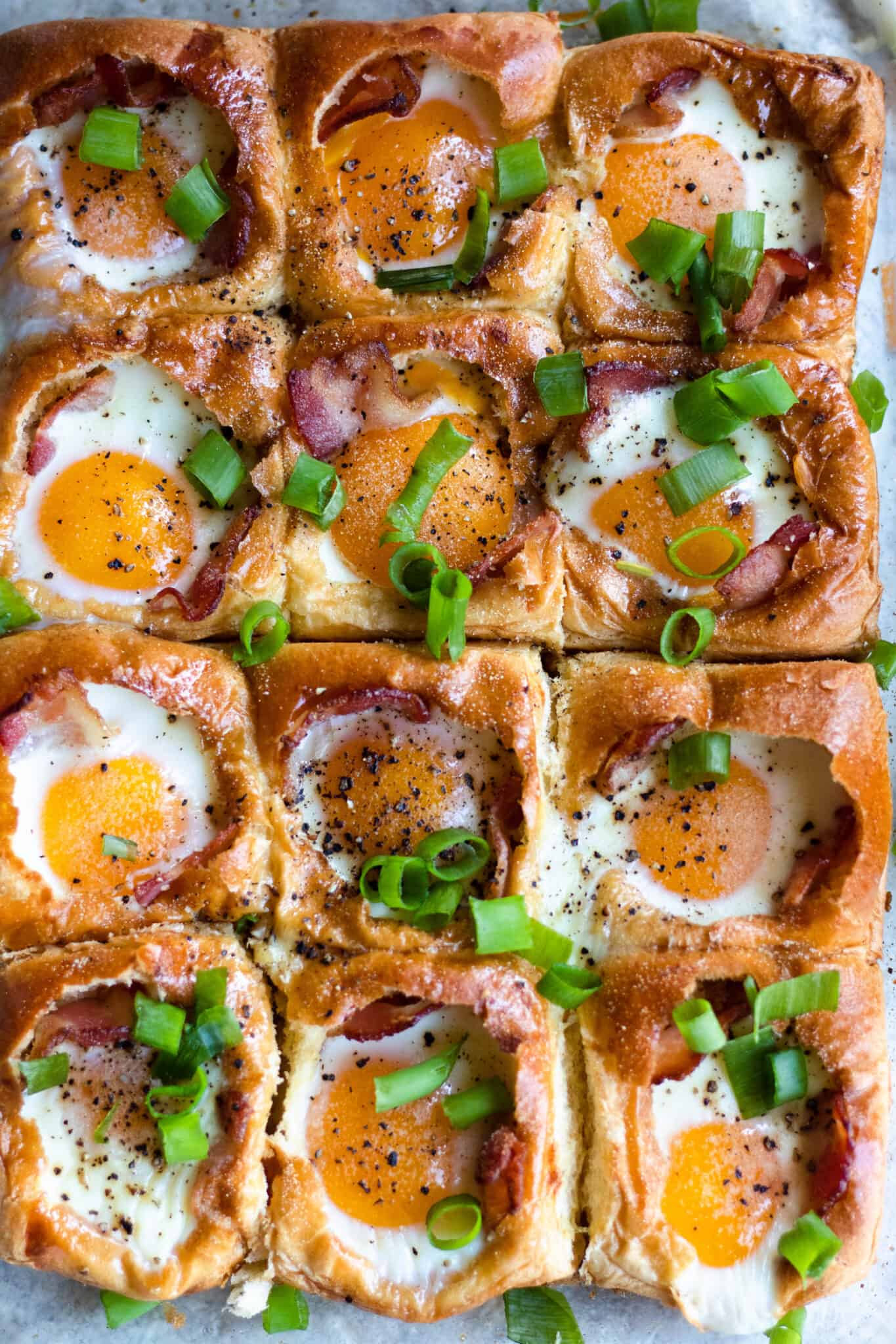 Nine squares of baked eggs with bacon on bread, topped with sliced green onions and black pepper.