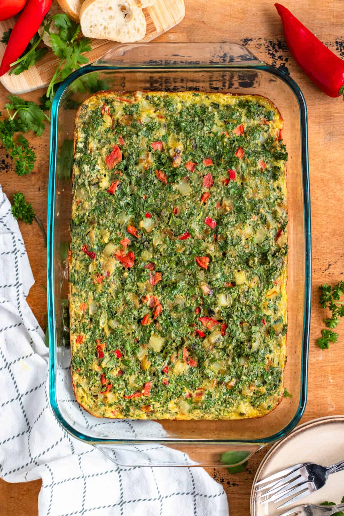A baked spinach and vegetable casserole in a glass dish on a wooden table, surrounded by herbs and bread.