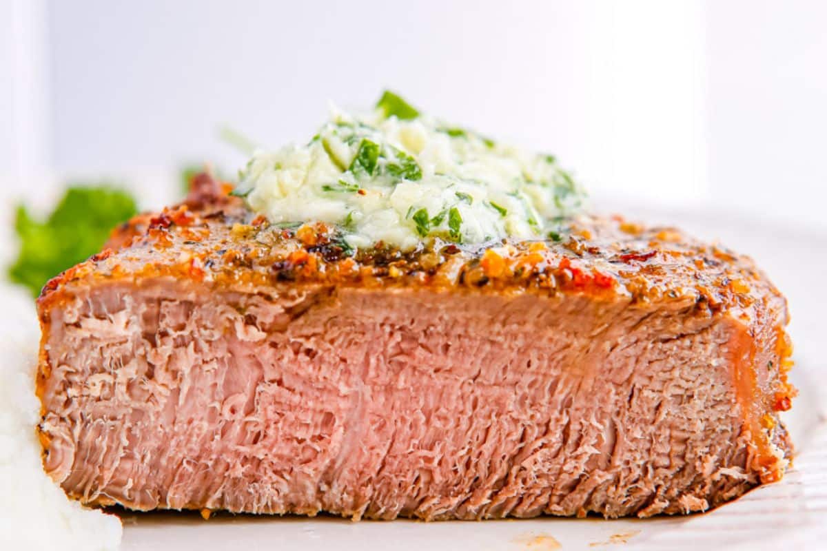 Close-up of a medium-cooked steak topped with herb butter, served with mashed potatoes on the side.