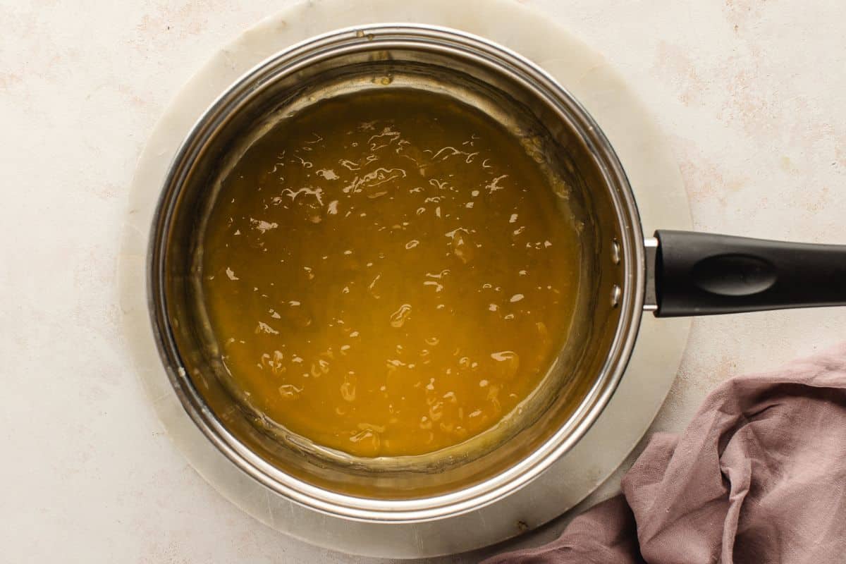 A saucepan filled with thick yellow lemon curd on a light surface, next to a mauve cloth.