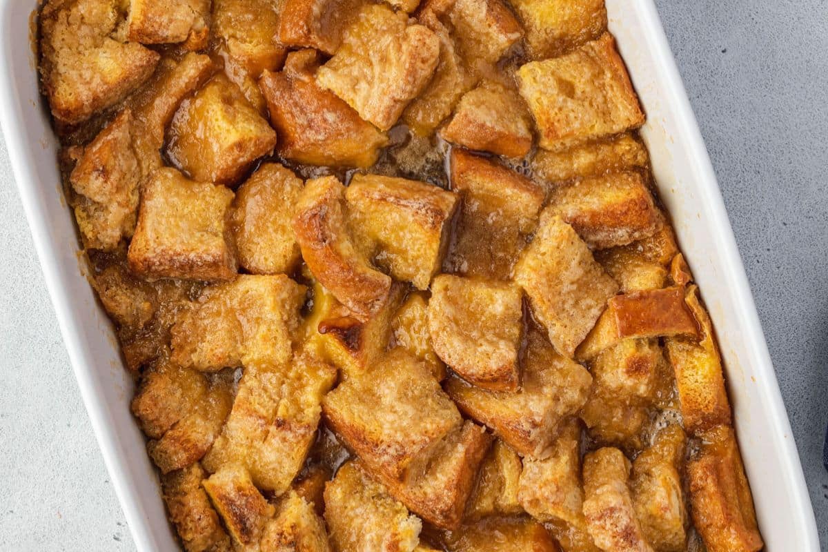 Close-up of golden-brown bread pudding in a white baking dish on a light gray surface.