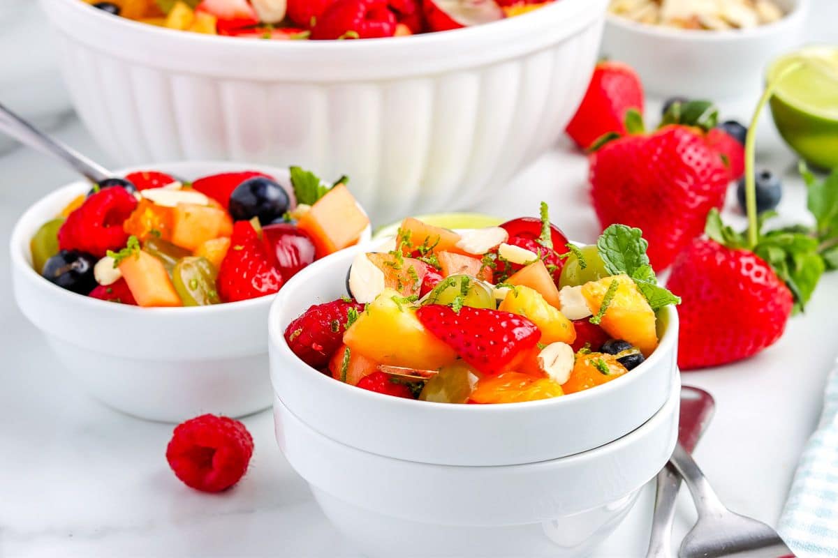 Colorful fruit salad with strawberries, blueberries, and nuts in white bowls, with fresh fruits around.