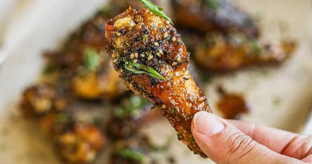 A hand holding a seasoned chicken wing topped with herbs, with more wings blurred in the background.
