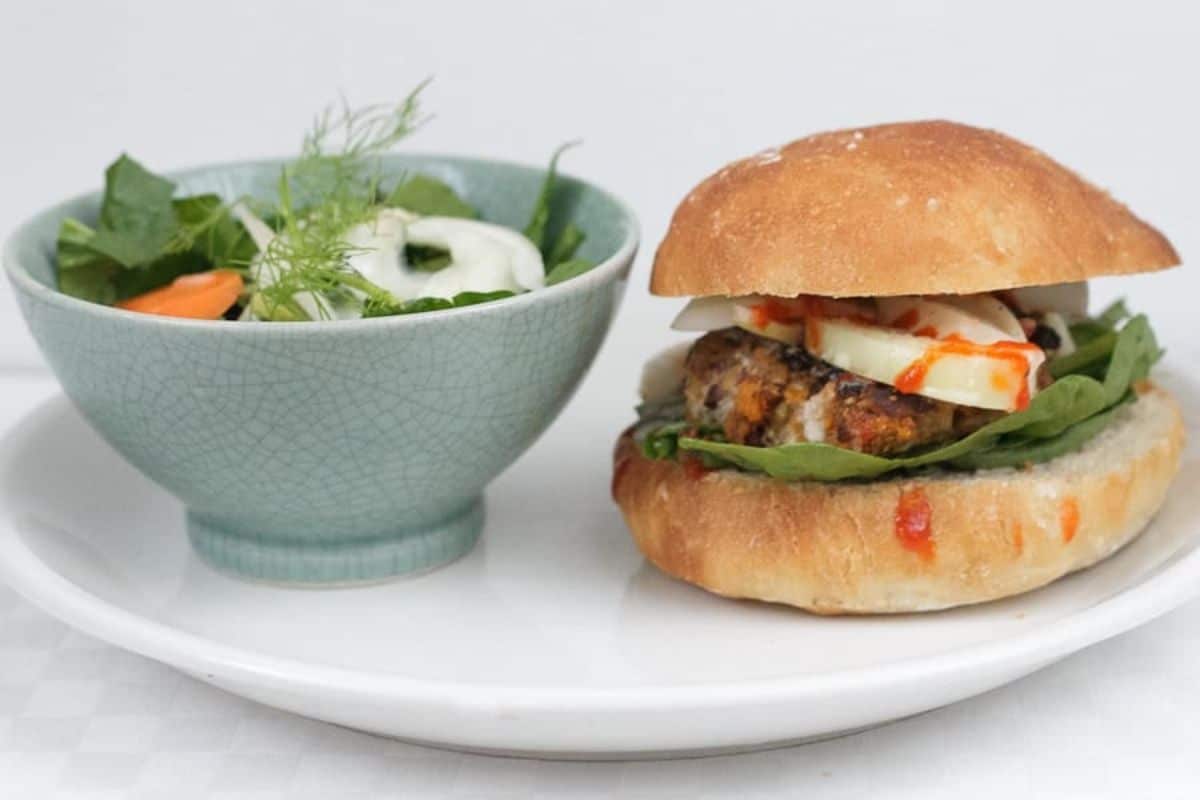 A burger with greens and cheese next to a bowl of salad on a white plate.